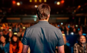 Man in a dark shirt on stage, facing away from the camera, with a blurred audience in the background.